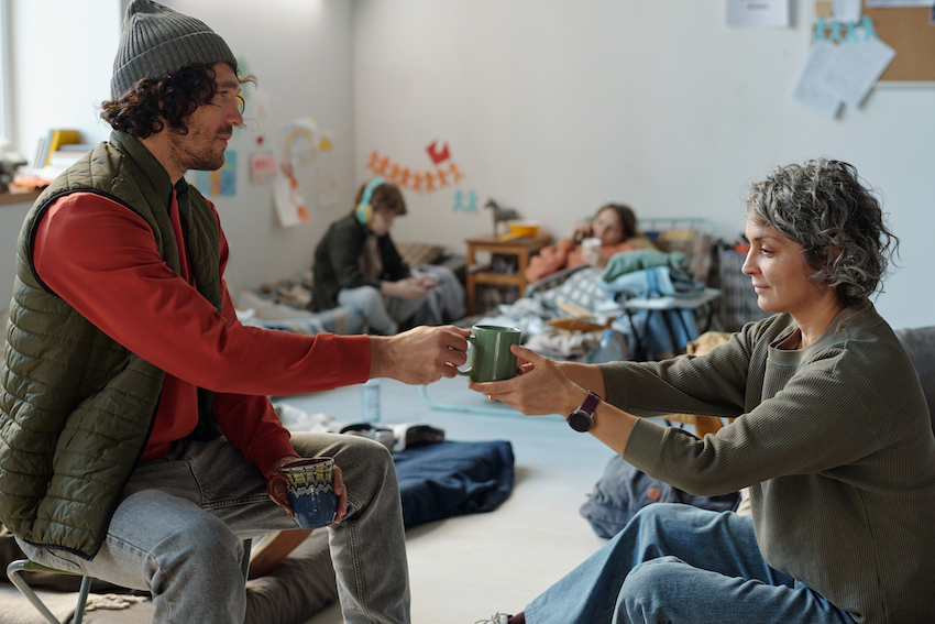 Mature woman taking cup of tea from hands of young male refugee while both sitting in front of camera in crisis camp